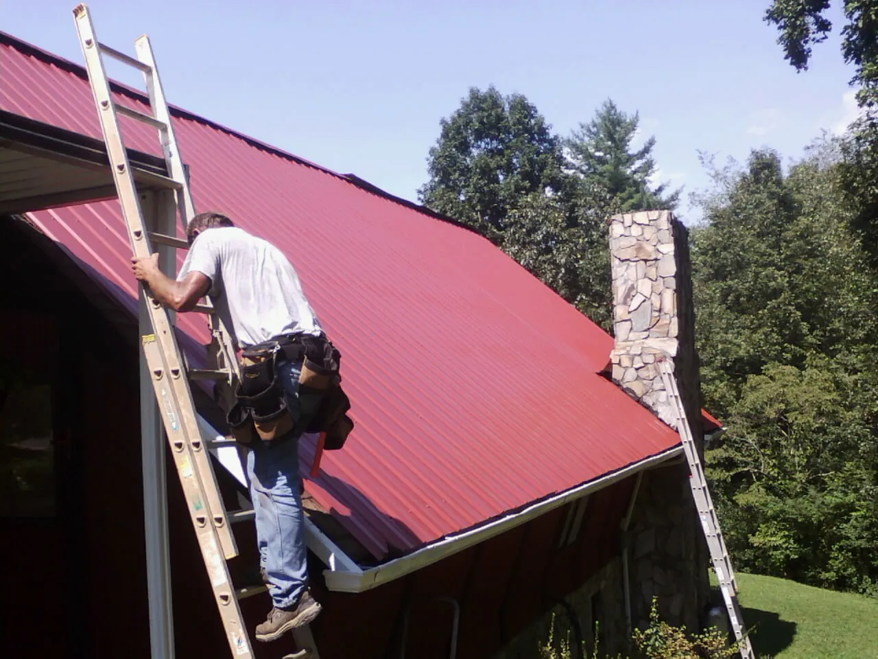 Roofer on a ladder beside a red metal roof and stone chimney — Soffit & Fascia Repair by Roofing Brothers in Alpharetta